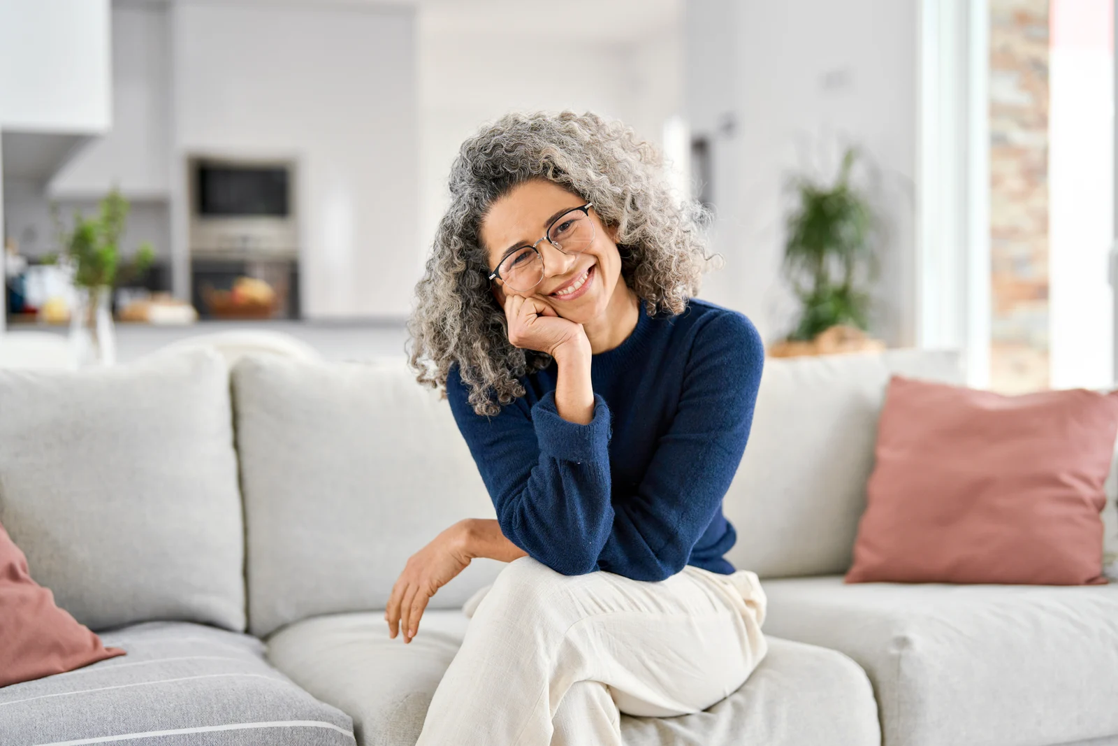 A smiling woman with gray curly hair and glasses seated on a sofa